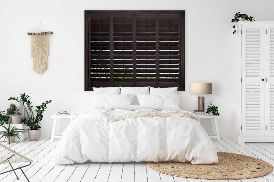Dark wood plantation shutters on a window over a bed in a white bedroom